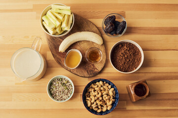 Flatlay of smoothie ingredients such as fresh peeled banana, cocoa powder, cashew nuts, milk, muesli and honey on wooden kitchen table