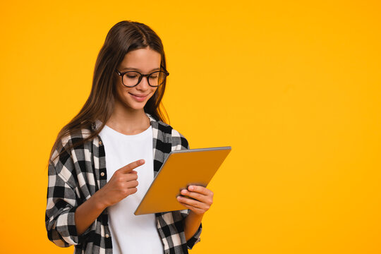 Smart young teenager girl schoolchild pupil student using digital tablet pointing at screen, surfing web pages social media, blogging, vlogging isolated in yellow background