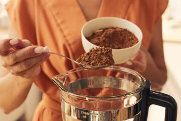 Hands of female putting spoon of cocoa powder into electric blender before mixing it with other...