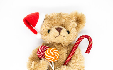 A stuffed toy Teddy bear in a red Santa Claus hat on one ear, holding candy cane and two striped lollipops in its paws. White background, copy space. Concept of Christmas