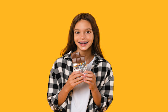 Young girl having sweet tooth. Caucasian teenager schoolgirl pupil child eating chocolate bar isolated in yellow background. Dental teeth problems, caries, children obesity concept.