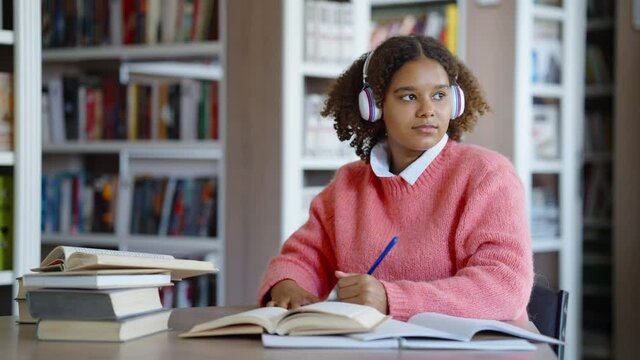 African American female student studying in library, stops writing, looking out window behind camera and listening to music in headphones, blurred background. Concept of education