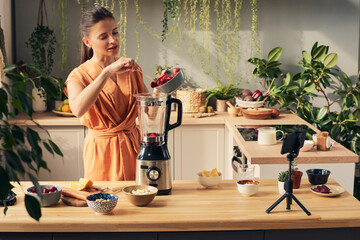 Young female blogger putting fresh strawberries into electric blender during masterclass about preparing homemade smoothie