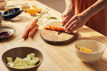 Hands of young housewife chopping fresh carrots for vegetable smoothie before putting them into electric blender