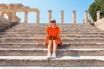 Young happy blonde woman sits on the steps of the Acropolis in the city of Lindos, Rhodes, Greece