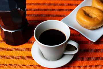 Cup of coffee next to a coffee pot with some glazed donuts on a white plate.