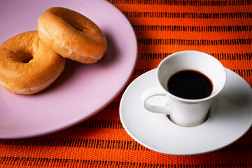 Breakfast or snack of a cup of coffee and some glazed donuts on an orange background.