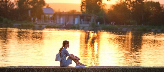Side view of lonely woman sitting alone on lake shore on warm evening. Solitude and relaxing in...