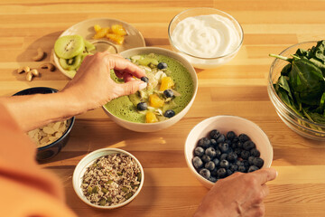 Young woman decorating green vegetables smoothie in bowl with fresh blueberries before serving it for breakfast