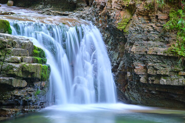 Obraz premium Waterfall on mountain river with white foamy water falling down from rocky formation in summer forest