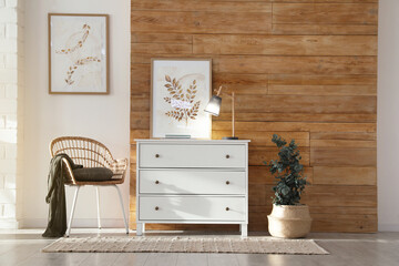 Stylish room interior with chest of drawers, wicker armchair and potted eucalyptus plant near wooden wall