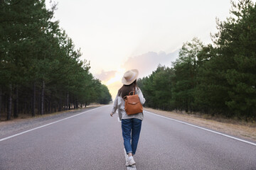 Young woman with stylish backpack walking along empty road, back view