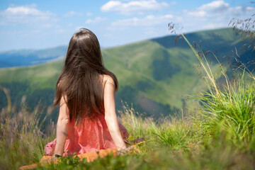 Young happy woman traveler in red dress resting on green grassy hillside on a windy day in summer mountains enjoying view of nature.
