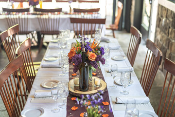 Table decorations at a wedding follow a purple and orange colored theme. The center pieces include flower petals, a wine bottle and purple sash going across the table.