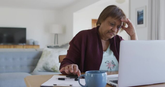 Worried african american senior woman at dining table, using laptop and holding head