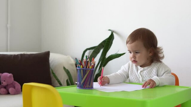 education caucasian little child daughter happy funny kid curly baby girl with draws picture at table. concentrated creative toddler writes with colored pencils on paper kids room. hobby study at home
