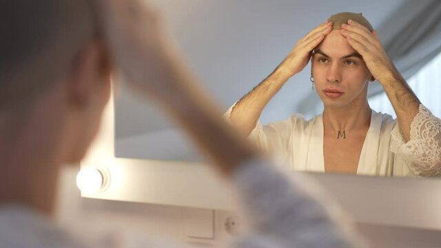 Confident queer woman putting on wig cap in slow motion reflecting in mirror. Reflection portrait of young handsome Caucasian non-binary person at home in bedroom in the morning