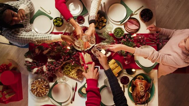 Friends celebrating new year top view, sitting at dining table. International young people pouring champagne into glasses and drinking, toasting. Diverse students during christmas party at home. 