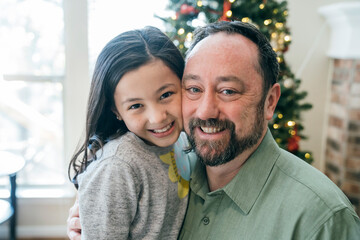 Portrait of happy girl hugging her smiling dad in front of Christmas tree 