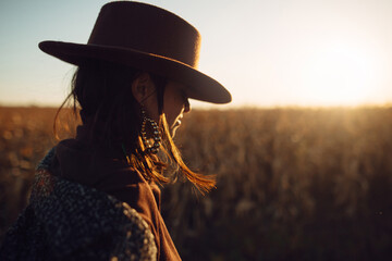 Beautiful stylish woman in hat walking in sunset light in autumn field. Atmospheric moment. Fashionable young hipster female in retro outfit enjoying evening at maize field in countryside
