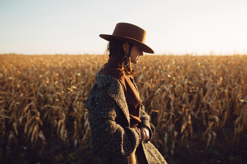Beautiful stylish woman in brown hat and vintage coat walking in sunset light in autumn field....