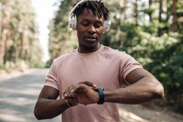 Fitness man in headphones who looks at his wristwatch while standing outdoors in the park