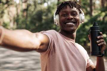 smiling african american man wearing headphones taking selfie after jogging holding water bottle...