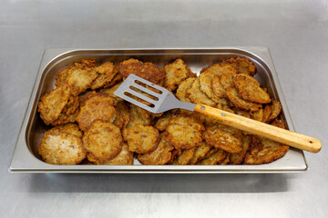 Lots of fried potato pancakes in a metal dish. On top is a spatula for frying.