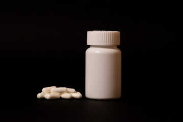 Many white capsules of sedatives, antivirals, vitamins lie near a white plastic jar on a black background