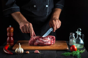 The cook cuts meat with a knife in the kitchen prepares food. Vegetables and spices on kitchen table in a restaurant to prepare a delicious lunch