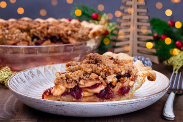 Slice of American apple cranberry pie, topped with crumbled dough and pecan, bokeh on background