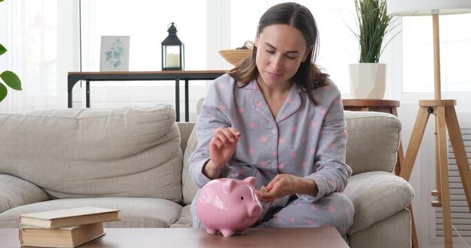 Happy woman inserting coin into piggy bank sitting on sofa at home