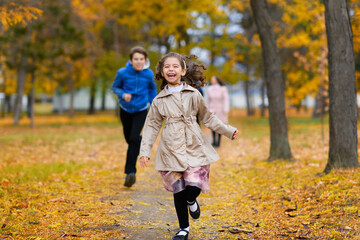 Fototapeta premium Children run along path in autumn park. They play and laugh, they have a lot of fun. Beautiful nature and trees with yellow leaves.