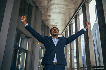 excited african american businessman rejoices in victory looking at camera while standing outside...