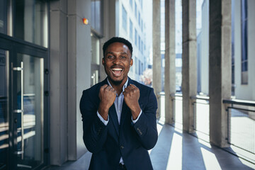 excited african american businessman rejoices in victory looking at camera while standing outside...