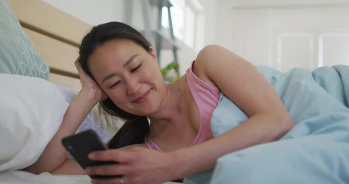 Happy asian woman lying in bed, using smartphone in the morning
