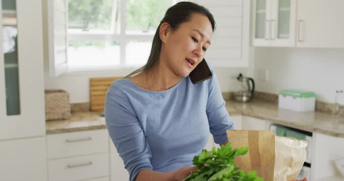 Happy Asian Woman Unpacking Groceries And Using Smartphone In Kitchen