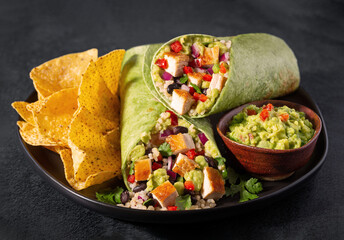 Chicken burrito in a spinach tortilla with yellow corn tortilla chips on a dark surface; copy space