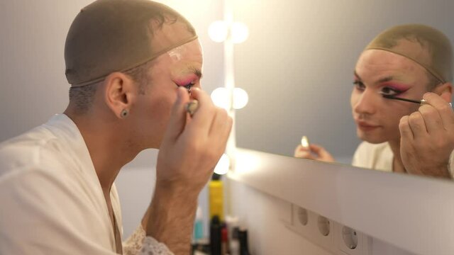 Side view focused gender-fluid woman applying mascara in slow motion reflecting in mirror. Concentrated proud confident Caucasian non-binary person in wig cap doing makeup at home. Lifestyle concept