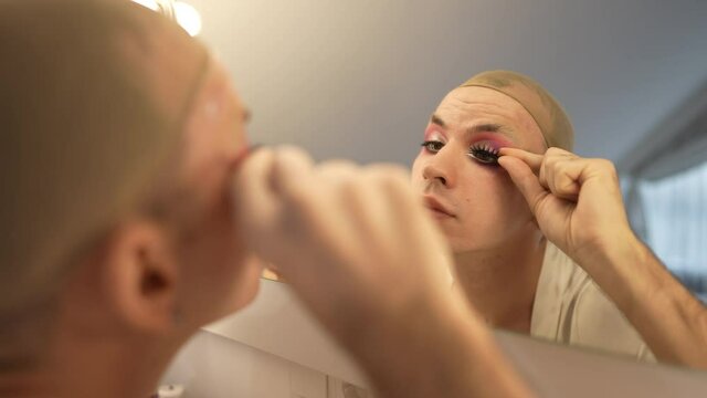 Concentrated gender-fluid woman applying false eyelashes in slow motion reflecting in mirror. Portrait of focused confident Caucasian millennial LGBT person doing makeup indoors. Style and diversity