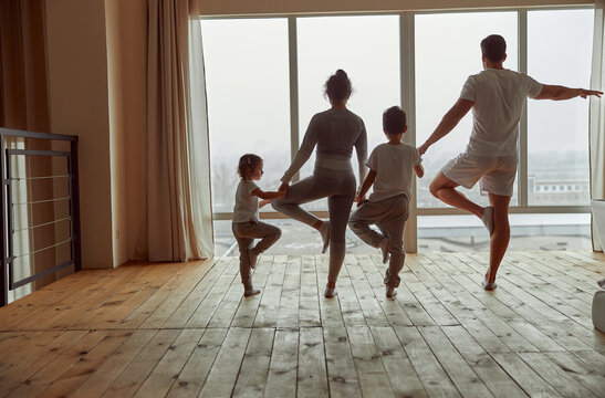 Sporty parents with son and daughter are standing on one foot in asna with faces to window