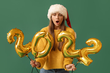 Shocked young woman in Santa hat with balloons in shape of figure 2022 on green background