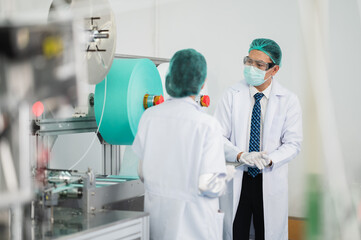 Fototapeta premium Both male and female Asian scientists are outfitted with protective gear. Examining the factory's machinery and materials used in the mask-making process. Covid 19 is a piece of preventive equipment.
