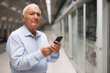 Obraz na płótnie Canvas European old man standing in subway station and using smartphone while waiting for train.