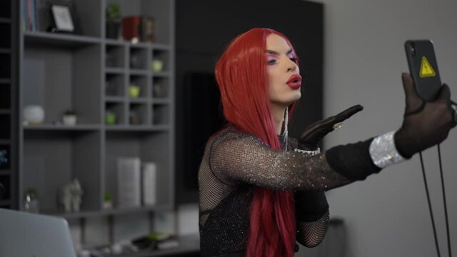 Portrait of coquette travesty woman sending air kiss blogging online sitting with smartphone indoors. Smiling confident Caucasian gender-fluid LGBT person streaming online from home in slow motion
