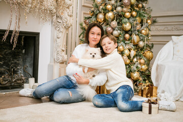 Happy family, mom and son. They are sitting near a christmas tree with a dog. Woman with family at christmas time at home. Happy hugging in sweaters caucasian family waiting for the new year