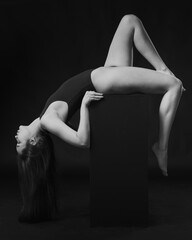 Pretty brunette woman in bodysuit posing on cube on black background. Black and white