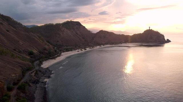 Rising over a secluded curved bay beach, hill landscape and Cristo Rei Jesus Christ statue on tropical island in Dili, Timor Leste. Aerial drone with stunning sunset tropical vibes.