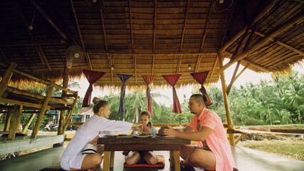 The happy family eat and drink tasty beverages spending time in local floating cafe on water. Father, mother and daughter having breakfast outdoors. Man, woman and a child in a cafe. Tropical view