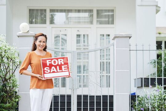 Smiling Mature Woman Selling Her House, She Is Standing At Gate With Sighboard